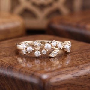 Op de afbeelding: Een gouden ring met een takvormig ontwerp, versierd met kleine witte parels en ronde, heldere stenen. De ringband is gemaakt van gepolijst geelgoud, waarbij de decoratieve elementen in een cluster aan de bovenzijde zijn geplaatst.