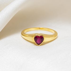 May include: A gold-colored ring with a heart-shaped, deep red gemstone. The ring is set on a white surface, and the band has a smooth, polished finish. The gemstone is a vibrant, eye-catching focal point.