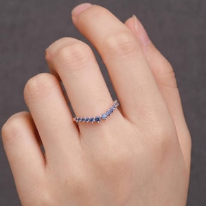 Pode incluir: Um anel em forma de V feito de uma banda metálica, apresentando uma série de pequenas pedras preciosas redondas de cor azul cravadas ao longo da curva superior. O design segue uma silhueta em chevron com um arco simétrico de pedras.