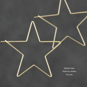 May include: Two gold star-shaped earrings. The earrings are made of thin wire and are very delicate. The text "Distant stars Hold my wishes For you" is written below the earrings.