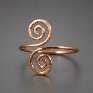 May include: A copper-colored metal ring featuring an open band design. The two ends of the band terminate in symmetrical, outward-facing spiral coils. The metal has a smooth, polished finish with a warm, reddish-brown metallic tone throughout.