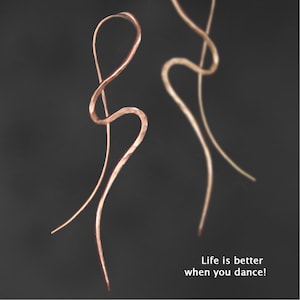 May include: A pair of long, slender, hammered copper-toned earrings with a fluid, curved, wire-like design. The text "Life is better when you dance!" is printed in white in the lower right corner against a dark, neutral background.