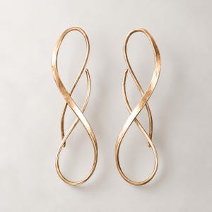 K&ouml;nnte beinhalten: Ein Paar lange, goldfarbene Ohrringe mit einem l&auml;nglichen, offenen Unendlichkeitsdesign. Die Metalloberfl&auml;che weist eine dezente, geh&auml;mmerte Textur auf, die das Licht einf&auml;ngt und eine flie&szlig;ende, durchgehende Form in einer Acht bildet.