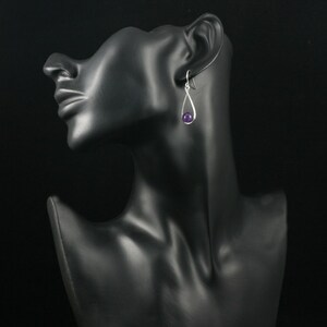 May include: Silver dangle earrings with a purple gemstone. The earrings are on a black mannequin head.