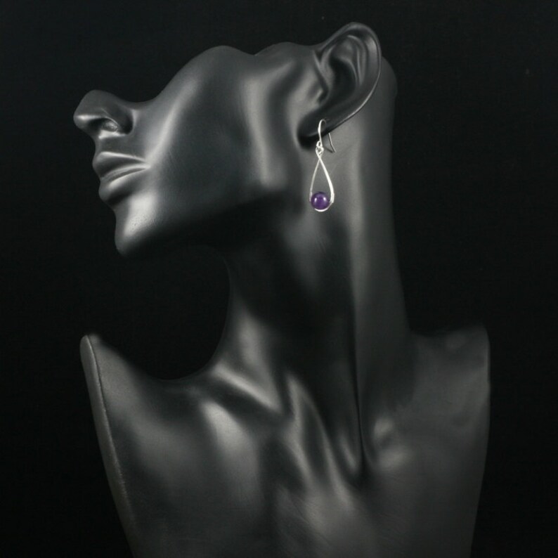 May include: Silver dangle earrings with a purple gemstone. The earrings are on a black mannequin head.
