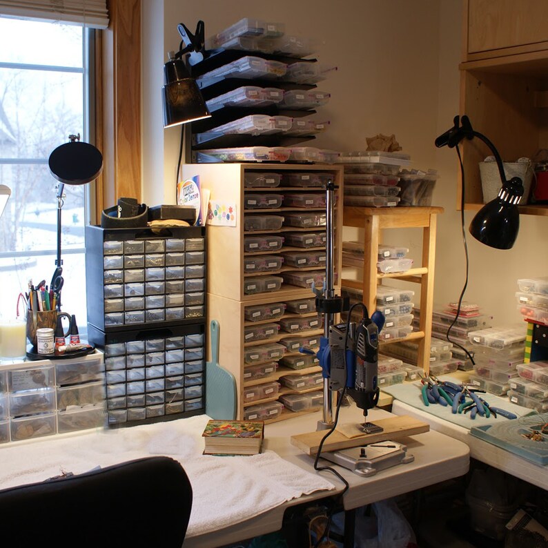 May include: A workspace with a variety of tools and supplies for jewelry making. There are multiple drawers filled with beads, a drill press, and a variety of tools on the table.