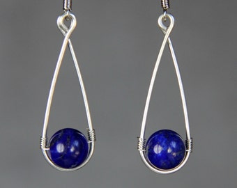 Op de afbeelding: Een paar zilveren oorhangers met een blauwe lapis lazuli-kraal die aan elke oorhanger hangt. De oorhangers zijn gemaakt van draad en hebben een traanvorm.