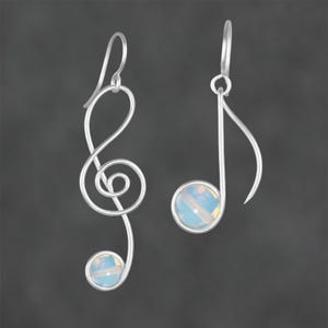 Op de afbeelding: Een paar oorbellen van zilverdraad in de vorm van muzieksymbolen. De ene oorbel heeft de vorm van een vioolsleutel met een ronde, iriserende cabochonsteen aan de onderkant. De andere oorbel is een muzieknoot met een bijpassende iriserende steen.