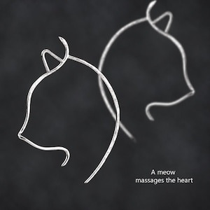 May include: Two silver-toned earrings shaped like the outline of a cat head with pointed ears. The metal has a textured, hammered finish. The text "A meow massages the heart" is printed in white in the lower right corner of the image.