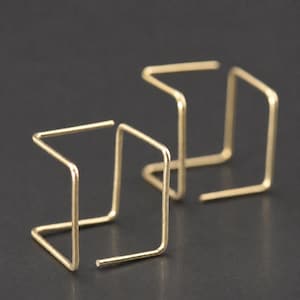 May include: A pair of gold-toned earrings featuring a geometric, open-frame square design. Each earring is constructed from a single thin metal wire bent into a three-dimensional, hollow cube shape with an open side, showcasing a clean, angular form.