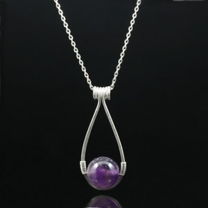 May include: A silver chain necklace with a teardrop-shaped pendant. The pendant holds a round, purple amethyst gemstone.