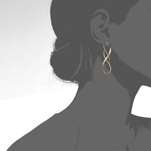 K&ouml;nnte beinhalten: Ein goldfarbener Ohrring aus einem schlanken, l&auml;nglichen Metalldraht, der sich zu einer durchgehenden, offenen Unendlichkeitsschleife windet. Das Design besteht aus einem einzelnen, glatten Metallband, das sich elegant zu einer feinen Form biegt.