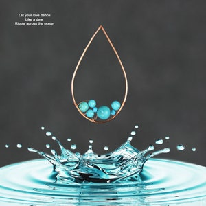 Pu&ograve; includere: Un orecchino a cerchio a forma di lacrima in filo di rame con perline blu turchese che pendono da esso. L'orecchino &egrave; sospeso sopra uno schizzo d'acqua blu.