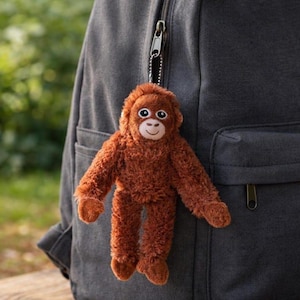 May include: A plush orangutan keyring with shaggy, rust-coloured fur, a light brown face, and dark eyes. The stuffed animal features long arms and legs, and it is attached to a silver-toned metal chain and clasp, hanging from a dark grey backpack.