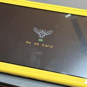 Puede incluir: Consola de juegos port&aacute;til amarilla con una pantalla negra que muestra un gr&aacute;fico de una frambuesa alada y el texto "No SD Card". Tambi&eacute;n se muestra un icono de bater&iacute;a.