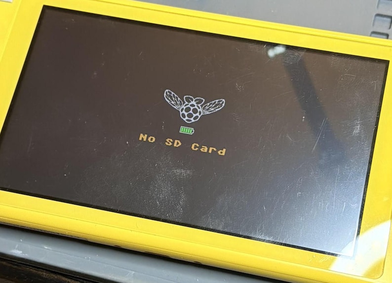 Puede incluir: Consola de juegos port&aacute;til amarilla con una pantalla negra que muestra un gr&aacute;fico de una frambuesa alada y el texto "No SD Card". Tambi&eacute;n se muestra un icono de bater&iacute;a.
