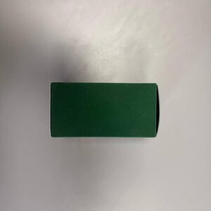 May include: A rectangular, dark green box with a matte finish. The box is positioned horizontally against a white background, showcasing its clean lines and simple design. The box appears to be empty.
