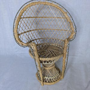 May include: A small, woven wicker peacock chair with a rounded back and a circular seat. The chair features a natural tan color with black and white trim along the edges. The chair is designed with a decorative, open weave pattern.