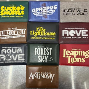 Puede incluir: Diez carteras rectangulares para tarjetas de varios colores, cada una con un título de juego de mesa y el nombre del autor impresos en el frente, incluyendo Cuckoo Shuffle, Apropos of Board Games, The Boy Who Cried Wolf, Lone Cowboy, The Last Lighthouse, Rove, Aqua Rove, Forest Sky, Leaping Lions y Antinomy.