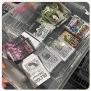 May include: A clear plastic storage bin containing multiple stacks of rectangular stickers and cards. The items feature various graphic designs, including black and white illustrations, colorful artwork, and text such as RUN, GLORIOUS, and HELL.