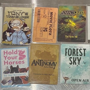 May include: A collection of ten rectangular game cards featuring various graphic designs, including titles like Tony's Treasures, AdvanceQuest, Skulls of Sedlec, Hold Your Horses, Antinomy, Forest Sky, and Arcane Bakery Clash, alongside a movie ticket.