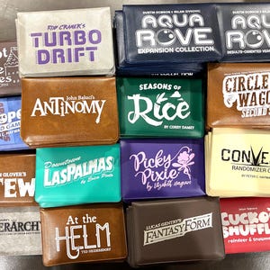 Puede incluir: Una colección de cajas rectangulares para juegos de cartas en varios colores, incluyendo marrón, azul, verde, morado y crema. Cada caja presenta títulos gráficos y texto impreso en blanco o morado, como Turbo Drift, Aqua Rove y Picky Pixie.