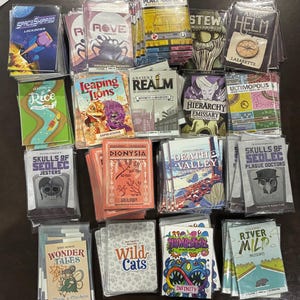 Könnte beinhalten: Eine Sammlung verschiedener Kartenspiele in durchsichtigen Kunststoffhüllen, darunter Titel wie SpaceShipped, Rove, Leaping Lions, Ancient Realm, Hierarchy Emissary, Skulls of Sedlec, Dionysia, Death Valley, Wonder Tales, Wild Cats, Numbsters und River Mild.
