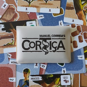 Pode incluir: Um jogo de tabuleiro intitulado "Corsica" de Manuel Correia, com uma caixa branca com o logotipo do jogo em preto. As cartas do jogo estão espalhadas ao redor da caixa, mostrando ilustrações e texto, incluindo as palavras "REST" e "PROCESS".