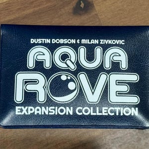 Op de afbeelding: Een marineblauwe spelcase met de woorden "AQUA ROVE" in grote witte letters. De tekst "EXPANSION COLLECTION" staat onder de hoofdtitel. De namen "DUSTIN DOBSON & MILAN ZIVKOVIC" staan boven de titel.