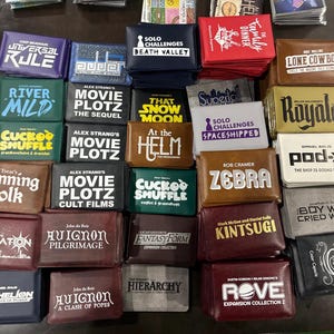 Puede incluir: Una colección de cajas de juegos rectangulares dispuestas en una cuadrícula, con varios títulos como Universal Rule, Death Valley, Movie Plotz, Cuckoo Shuffle, Cunning Folk, Avignon, Anthelion, Lone Cowboy, Royale, Pod-X, Zebra, Kintsugi y Rove.