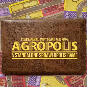 May include: A brown board game box with the title "AGROPOLIS" in yellow lettering. The game is for 1-4 players. The background features a colourful game board design with roads and fields.