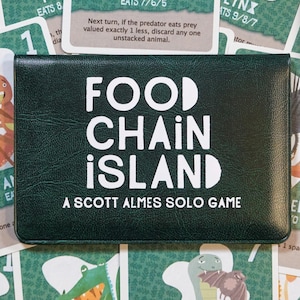 Op de afbeelding: Een donkergroene speldoos met de titel "FOOD CHAIN ISLAND" in witte letters. Het spel is een solo-spel van Scott Almes. De afbeelding bevat speelkaarten met dierenillustraties en de tekst "PLAYERS 1".