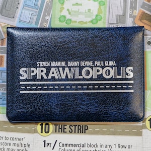 Może przedstawiać: Ciemnoniebieskie pudełko gry planszowej Sprawlopolis z białym tekstem i projektem drogi. Gra jest dla 1-4 graczy. Pudełko znajduje się na kartach do gry z mapami miast i instrukcjami.