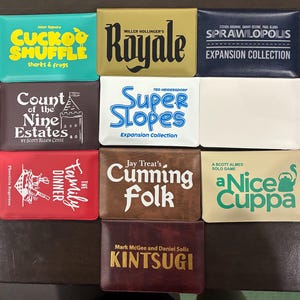 May include: A collection of ten rectangular card wallets featuring various game titles and graphics, including Cuckoo Shuffle, Royale, Sprawlopolis, Count of the Nine Estates, Super Slopes, The Family Dinner, Cunning Folk, A Nice Cuppa, and Kintsugi.
