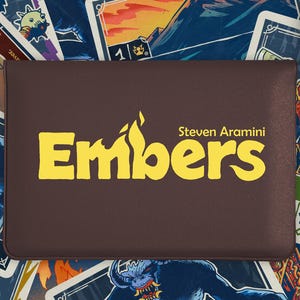 May include: A brown card game box with the word "Embers" in yellow, with the name Steven Aramini. The box is surrounded by illustrated cards with fantasy creatures. The top right corner shows "Players 1".