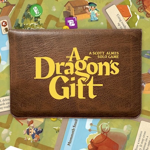 May include: A brown board game box with the title "A Dragon's Gift" in yellow lettering. The box is on a game board with cards and game pieces. The game is a solo game.