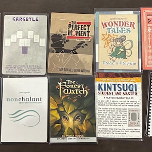 May include: A collection of ten rectangular game cards and rule sheets arranged in two rows. Titles include Gift Bow, Gargoyle, The Perfect Moment, Wonder Tales, Dionysia, Six Sons of the Sultan, Nonchalant, The Forest Watch, and Kintsugi.