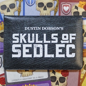 May include: A board game box titled "Skulls of Sedlec" in white, bold lettering on a black, textured surface. The game is for 2-3 players. Cards with skull illustrations and point values are visible. The game is by Dustin Dobson.