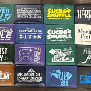 Puede incluir: Una colección de dieciséis carteras de juego rectangulares de varios colores, cada una con títulos y nombres de autor impresos en blanco o azul, incluyendo River Mild, Sprawlopolis, Cuckoo Shuffle, Hórreos, Universal Rule, Pentaquark y otros.