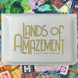 May include: A white game box with gold lettering that reads "LANDS OF AMAZEMENT" and "A SCOTT ALMES SOLO GAME." The background features colourful illustrations of amusement park rides and game cards. The game is for 1 player.