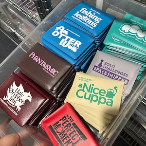Op de afbeelding: Een verzameling kleine, rechthoekige bordspeldozen in een doorzichtige plastic bak. Titels zijn onder andere Fishing Lessons, An Otter Won, Phantasmic, Solo Challenges Spaceshipped, Ugly Gryphon Inn, aNice Cuppa en The Perfect Moment in diverse kleuren.