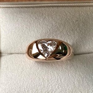 Pode incluir: Um anel de ouro rosa com um diamante em forma de coração em uma moldura. O anel é exibido em uma caixa cinza clara. O diamante brilha, refletindo a luz. A banda do anel é lisa e polida.