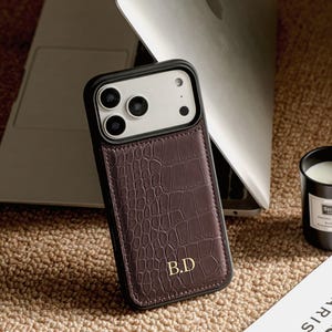 May include: A dark brown phone case with a textured crocodile skin pattern and a black frame. The case features the gold embossed initials B.D. at the bottom centre. The camera module area is white with three circular lens cutouts and a flash.