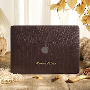 May include: A dark brown laptop cover with a textured crocodile skin pattern, featuring a central silver Apple logo cutout and the name Marian Oliver engraved in gold script lettering centered near the bottom edge of the rectangular case.