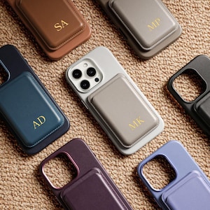 May include: A collection of personalized phone cases in various colors, including brown, gray, blue, and purple. Each case features a rectangular cardholder with gold initials. The cases are arranged on a textured, tan surface.