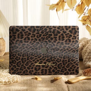 May include: A laptop skin featuring a brown and black leopard print pattern with a centered Apple logo cutout and the name Marian Oliver printed in a gold script font on the lower portion of the textured surface.