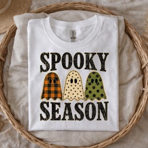 May include: A white short-sleeve t-shirt featuring the black text SPOOKY SEASON. Between the words are three ghost illustrations: one with an orange and black buffalo plaid pattern, one with a cream star pattern, and one with a green polka dot pattern.