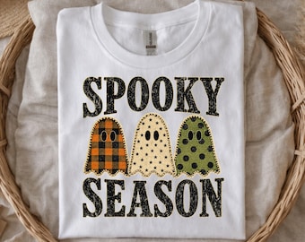 May include: A white short-sleeve t-shirt featuring the black text SPOOKY SEASON. Between the words are three ghost illustrations: one with an orange and black buffalo plaid pattern, one with a cream star pattern, and one with a green polka dot pattern.