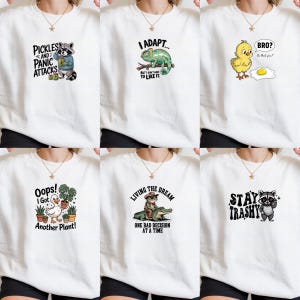 May include: Six white crewneck sweatshirts, each featuring a unique graphic print: a raccoon with pickles, a chameleon, a chick looking at an egg, a duck with potted plants, a frog riding an alligator, and a raccoon with the text Stay Trashy.