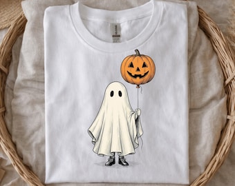May include: A white short-sleeved t-shirt featuring a graphic illustration of a ghost draped in a white sheet, wearing black shoes, and holding a string attached to an orange jack-o'-lantern pumpkin balloon with a carved face.
